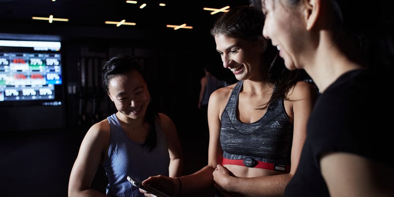 Three women looking at the Myzone app, with a Myzone display screen in the background