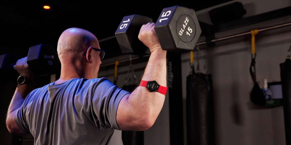 Man lifting two dumbbells in a workout studio, wearing a heart rate monitor