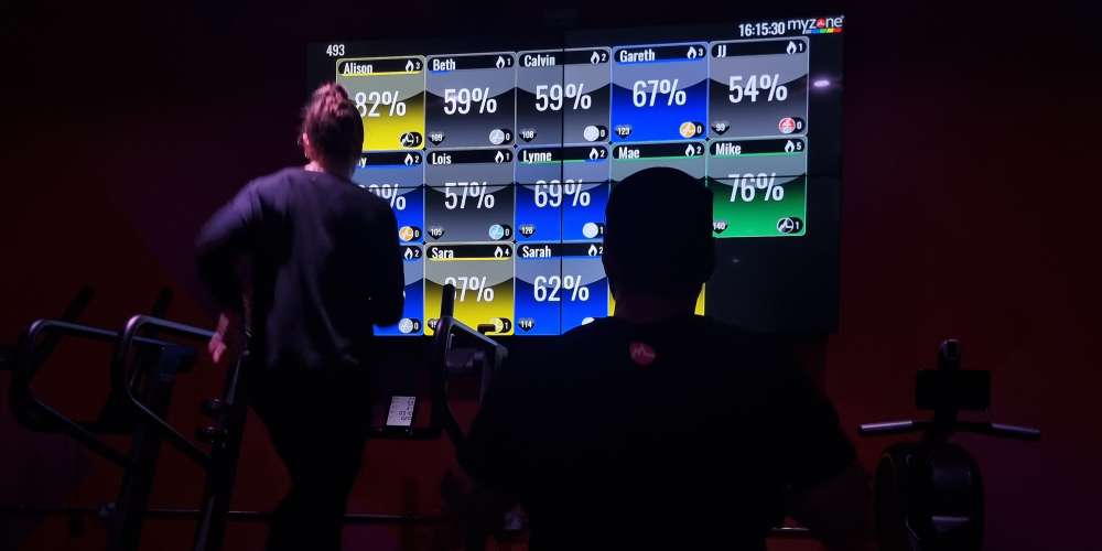 A man and a woman working out on cardio machines in the gym, in front of heart rate zone tiles on a screen