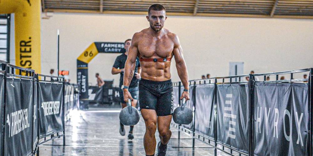 Jake Dearden performing a farmer's walk kettlebell carry at a HYROX event