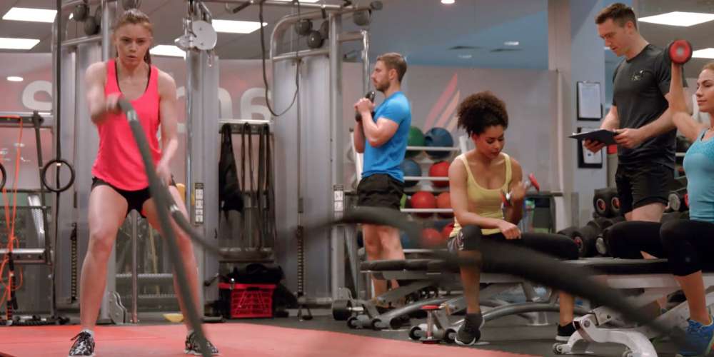 Men and women working out in the gym, watched by a personal trainer